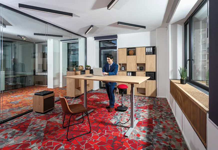 Muž pracující na notebooku u výškově stavitelného stolu v moderní kanceláři s designovým červeným kobercem a prosklenou stěnou.