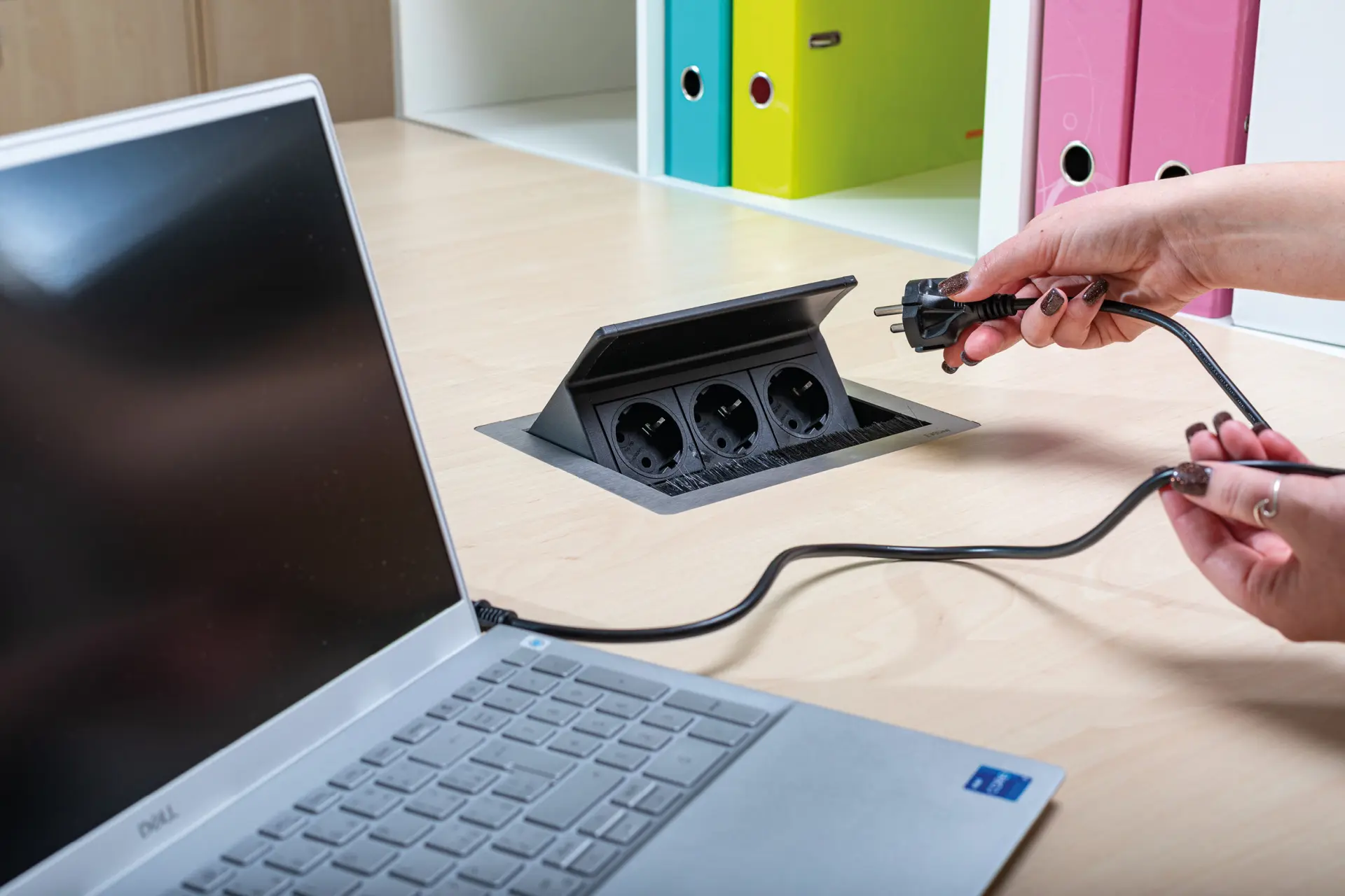 Detailní záběr na ruce zapojující černý kabel do výsuvné zásuvkové lišty zabudované v desce pracovního stolu. Vedle leží otevřený notebook.