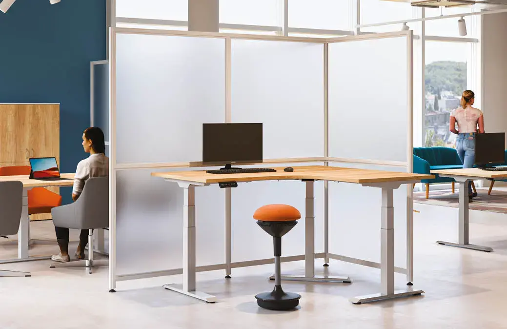 Moderní kancelářské pracoviště s rohovým výškově stavitelným stolem, ergonomickým oranžovým taburetem a poloprůhlednými paravány pro zajištění soukromí v otevřeném kancelářském prostoru.